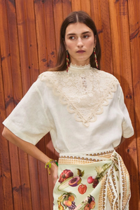Woman in a white lace top and colorful embroidered skirt standing against a wooden wall.